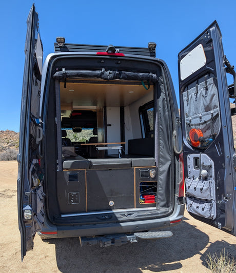 Rear view of an rv rental camper van with open doors, showcasing its custom interior, bed, table, and clever storage.