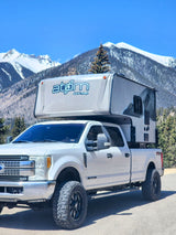Experience mountain adventures with this RV rental: a white Ford pickup truck carrying an atøm truck camper against snowy peaks.