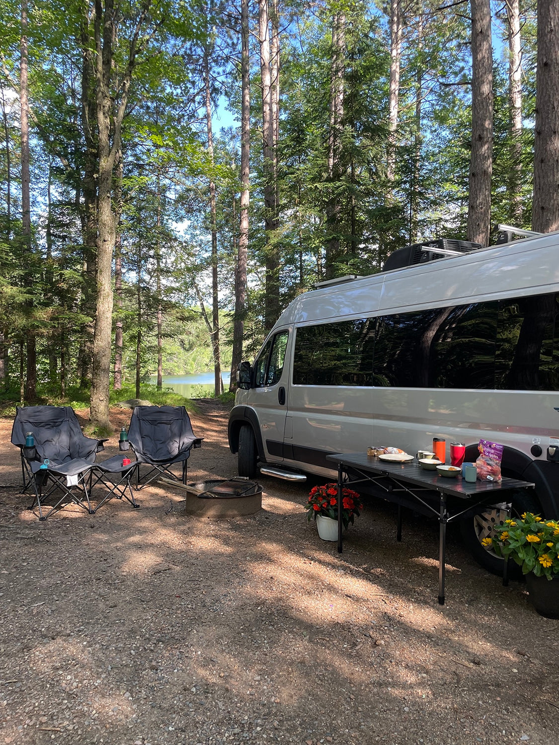 This RV rental offers a perfect camping setup: a van parked by a lake, with chairs, fire pit, and food on a table under tall trees.