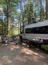 This RV rental offers a perfect camping setup: a van parked by a lake, with chairs, fire pit, and food on a table under tall trees.