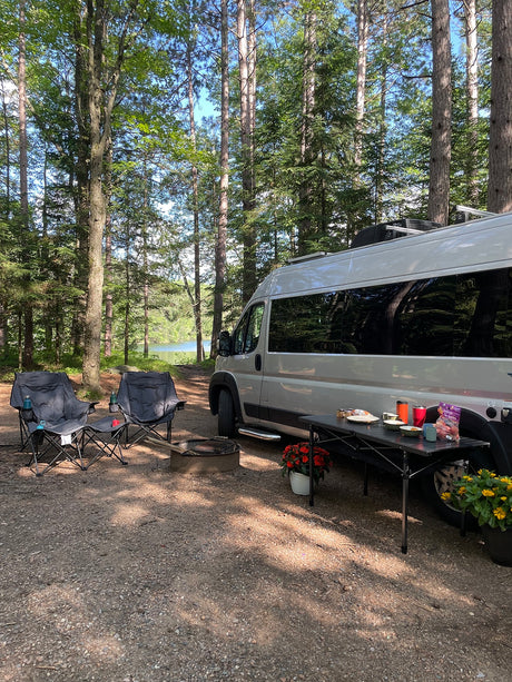 This RV rental offers a perfect camping setup: a van parked by a lake, with chairs, fire pit, and food on a table under tall trees.