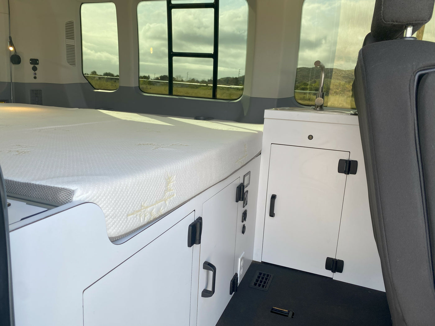 This RV rental interior features a cozy bed on the left, white cabinets with a sink, and window views for a comfortable journey. — photo 16