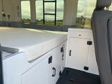 This RV rental interior features a cozy bed on the left, white cabinets with a sink, and window views for a comfortable journey.