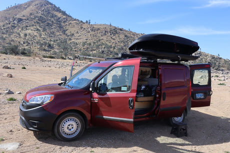 Red converted RAM cargo van (like an RV rental) with open doors, parked in rocky, mountainous terrain under a blue sky.