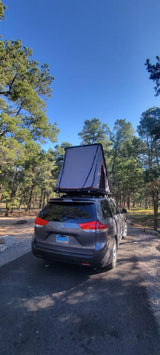 Explore the outdoors! Grey minivan with rooftop tent set up in a sunny pine forest. A flexible alternative to an RV rental.
