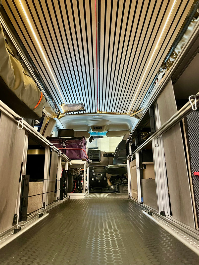  parked on asphalt amidst tall evergreen trees in a sun-dappled forest.|Step inside this modern **rv rental** camper van with modular storage — photo 2