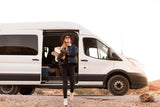 A woman exits a white **RV rental** van with firewood, dressed for camping in a vast desert landscape.