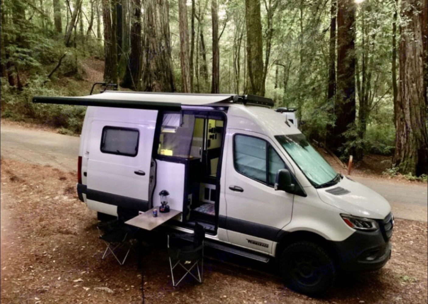RV rental camper van parked among towering redwoods. Awning out, table & chair ready for a perfect outdoor adventure.