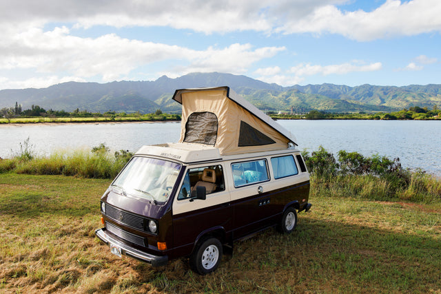 Brown & beige VW Westfalia pop-top camper van (RV rental) by a lake with scenic mountains. Ready for adventure!
