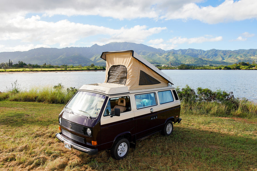 Brown & beige VW Westfalia pop-top camper van (RV rental) by a lake with scenic mountains. Ready for adventure!