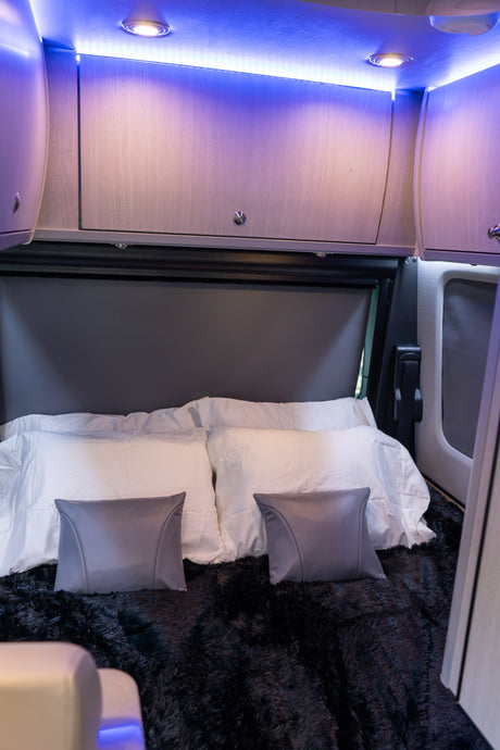 The bed area of an RV rental, featuring blue ambient lighting, overhead cabinets, white pillows, and a fluffy black blanket.