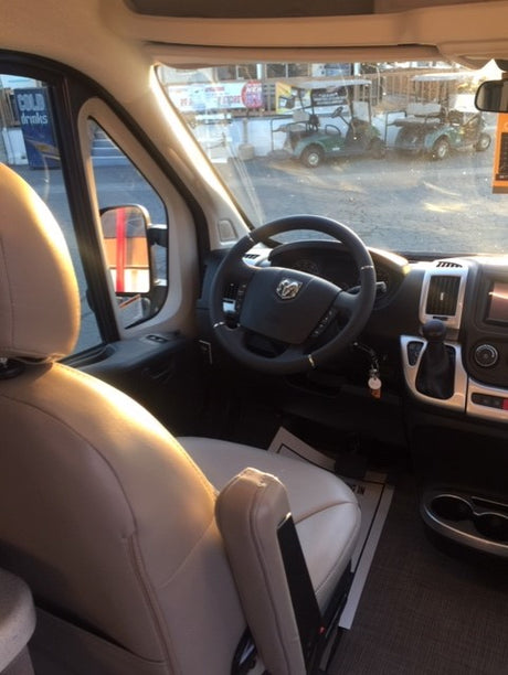 Driver's perspective inside an RV rental: Ram ProMaster cab with dashboard, seat, and steering wheel. Golf carts visible outside.