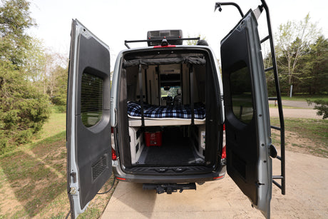 Rear view of a gray camper van RV rental, doors open revealing a comfy bed, storage, and roof access ladder.