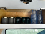 Inside an RV rental kitchen cabinet: neatly organized gray mugs, dark green glasses, and textured blue cups.