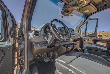 Interior view of an RV rental van's driver cockpit, featuring a modern dashboard and open door to scenic mountains.