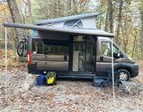 A grey pop-top RV rental with an extended Thule awning is set up in a leafy forest. A bike, chair, and table are visible.