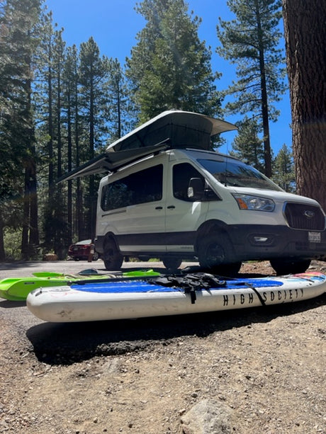 White RV rental with awning, paddleboard, & kayak parked in a sunny forest setting. Adventure awaits!