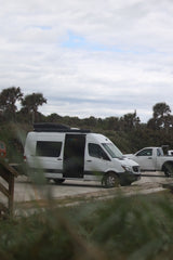 White RV rental Sprinter van with open side door, parked by palm trees under cloudy sky, perfect for a road trip.