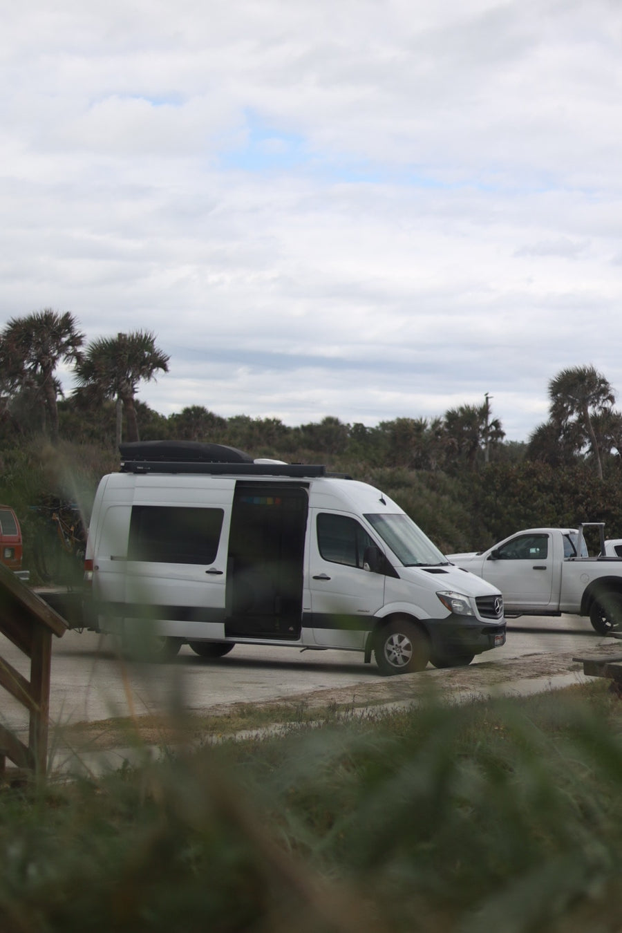 White RV rental Sprinter van with open side door, parked by palm trees under cloudy sky, perfect for a road trip.