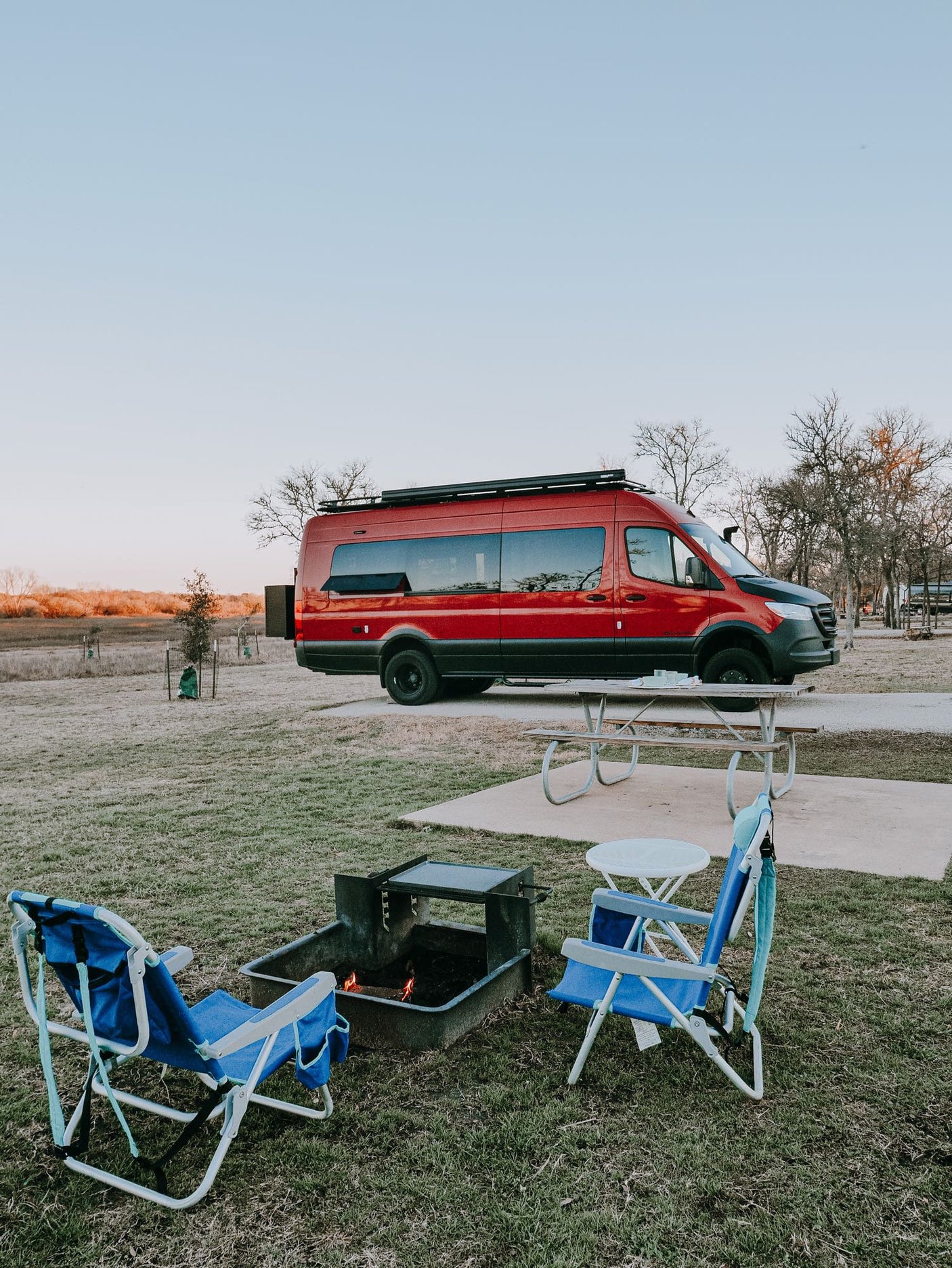 This red rv rental camper van is set up at a campsite with a lit fire pit and blue chairs for a relaxing evening.