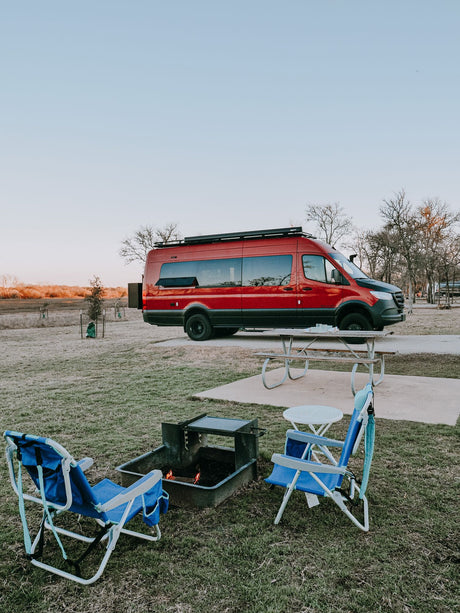 This red rv rental camper van is set up at a campsite with a lit fire pit and blue chairs for a relaxing evening.