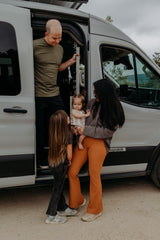Family by their rv rental camper van. Dad in door, mom holds baby, older child watches. Fun family travel.