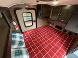 Cozy RV rental interior: a bed with red plaid bedding, overhead cabinets, an exit door with a window, and a fire extinguisher.
