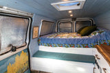 Cozy RV rental interior: custom-built bed, reflective window covers, soft blue paneling, and a small bench seat.