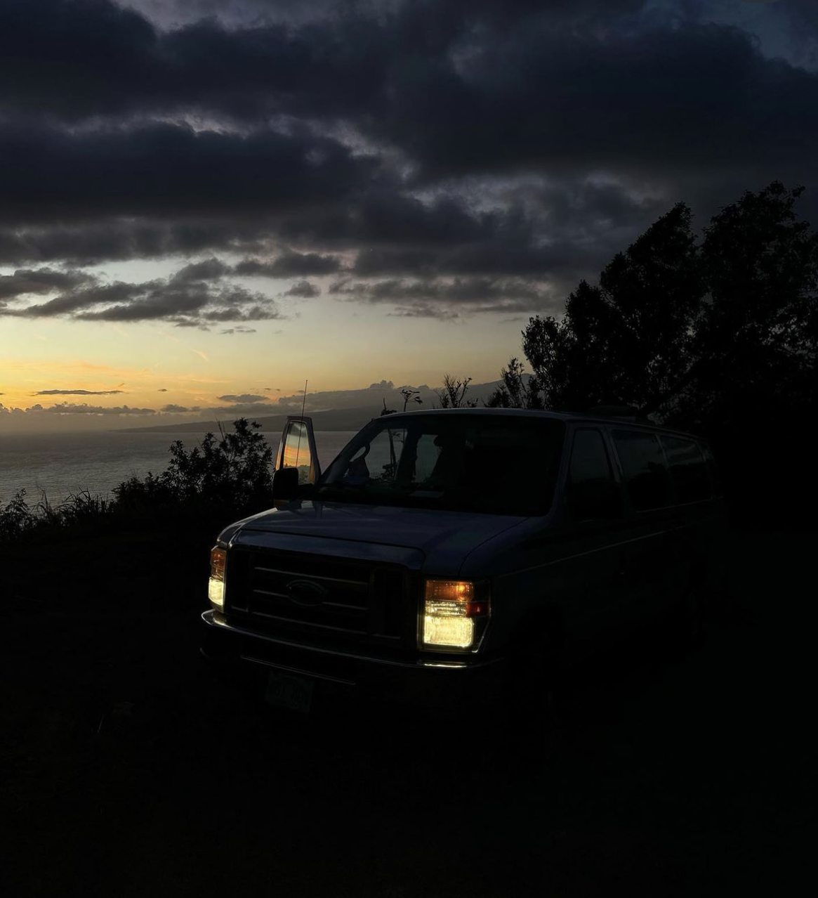 Silhouetted van with headlights on at sunset by ocean. Door open, reflecting sky. An RV rental adventure awaits!