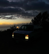 Silhouetted van with headlights on at sunset by ocean. Door open, reflecting sky. An RV rental adventure awaits!