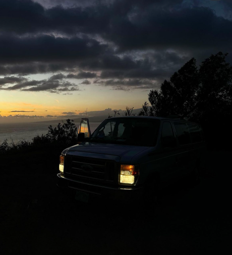 Silhouetted van with headlights on at sunset by ocean. Door open, reflecting sky. An RV rental adventure awaits!