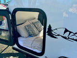 Comfort awaits in this RV rental! Open door shows a cozy bed inside this white travel trailer with a mountain graphic.