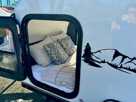 Comfort awaits in this RV rental! Open door shows a cozy bed inside this white travel trailer with a mountain graphic.