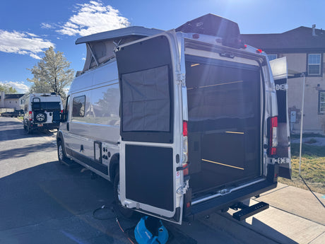 White camper van RV rental with open rear doors, pop-top roof, and AC unit. A second similar van is parked behind it.