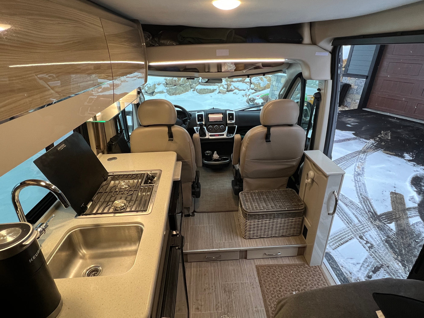 Inside a well-equipped RV rental: modern kitchen with sink & stove, dual cockpit seats, and snowy views outside. Great for travel.