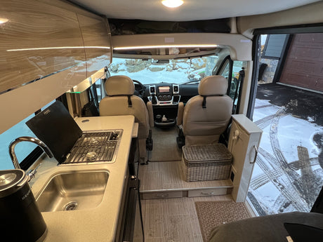 Inside a well-equipped RV rental: modern kitchen with sink & stove, dual cockpit seats, and snowy views outside. Great for travel.