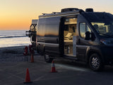An RV rental camper van parked by the ocean at sunset, its lighted interior visible through the open side door on a beach.