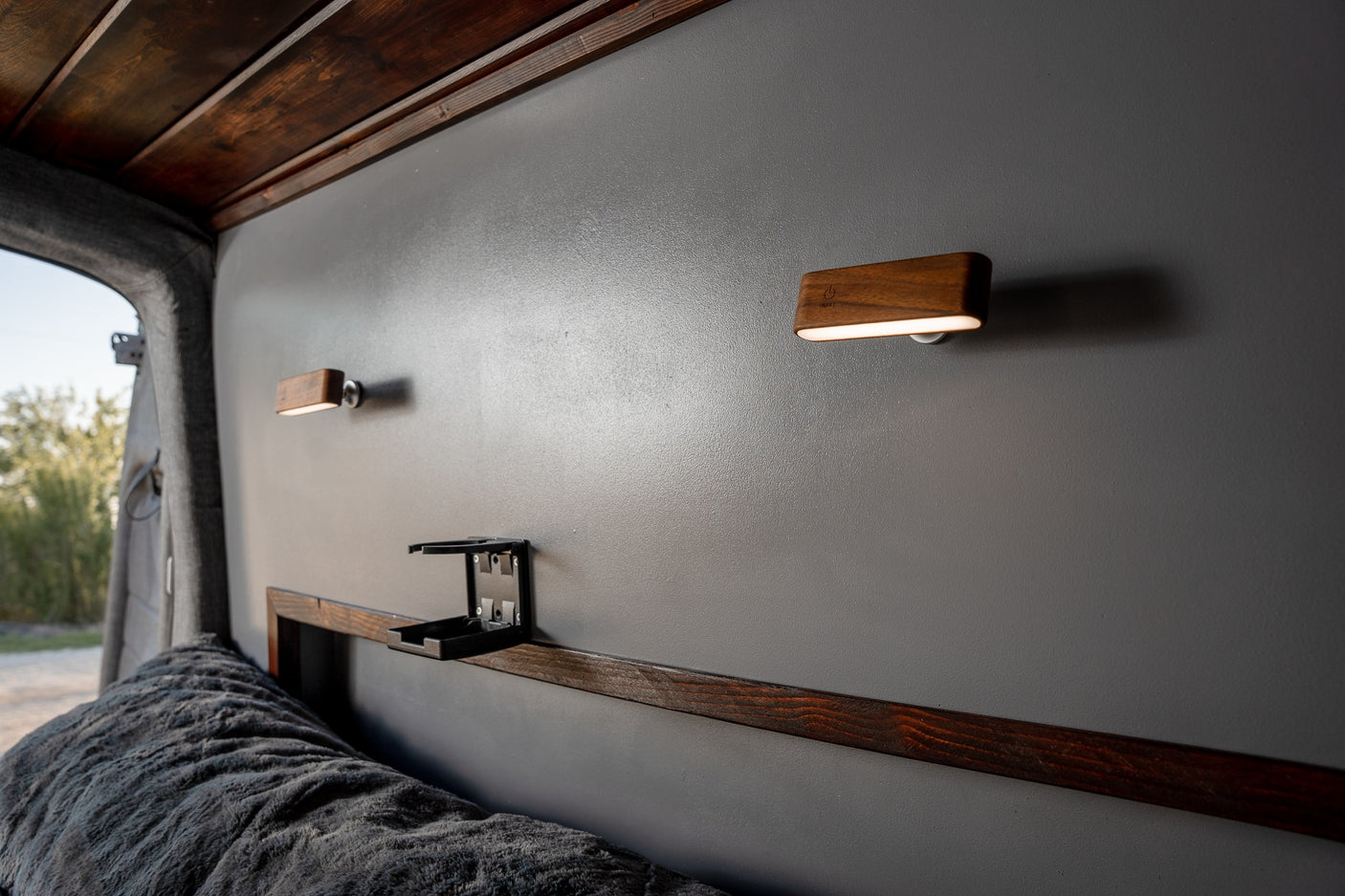 RV rental interior showing modern wooden wall lights, a foldable cup holder, and a cozy bed with dark wood trim.