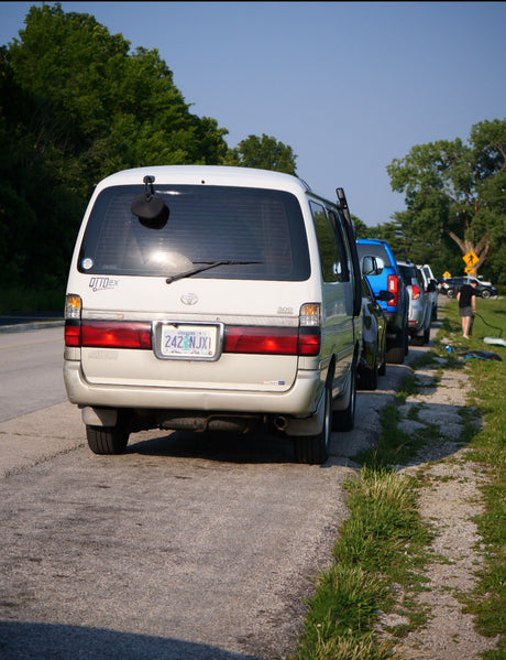 A white Toyota van, an RV rental option, parked on a sunny, tree-lined road with other vehicles.