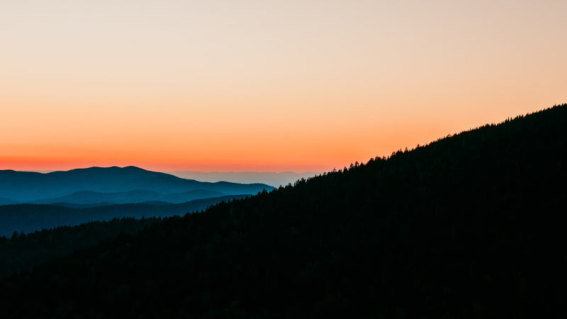 Vibrant orange sunset illuminates distant blue mountain layers & dark foreground trees. Imagine this view from an RV rental. — photo 13