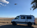 RV rental adventure awaits! A silver camper van is parked in a dry field under a blue sky with a large plane flying overhead.