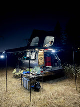 Night car camping setup: An SUV with a rooftop tent and awning provides an RV rental-like experience. Illuminated outdoor kitchen & relaxing space.