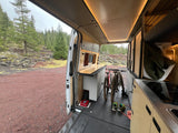 RV rental interior: two mountain bikes stored in a custom camper van, rear doors open to a pine forest and red earth.