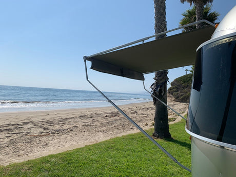 This RV rental offers a stunning beach view. An Airstream with awning extended, overlooking the ocean, sand, and palm trees.