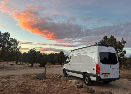 White Sprinter van (an RV rental) parked at sunset in a desert campsite, featuring colorful clouds. Ideal for outdoor adventures.
