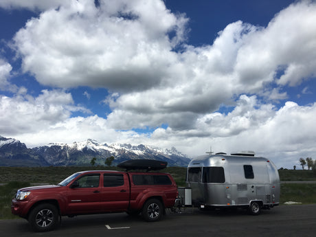 A red pickup truck tows a silver Airstream RV rental in front of majestic snowy mountains and a cloudy blue sky.