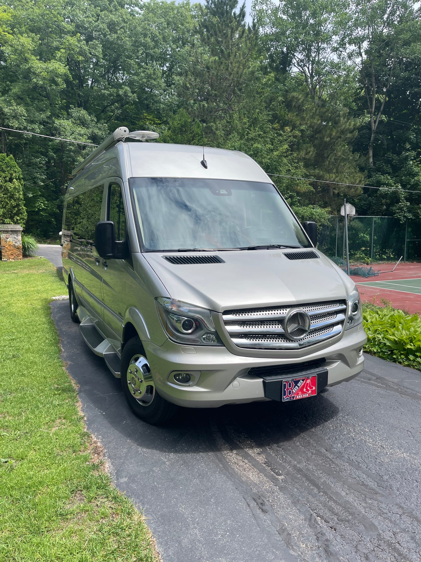 Silver Mercedes-Benz Sprinter RV rental parked on an asphalt driveway with trees and a tennis court in the background.