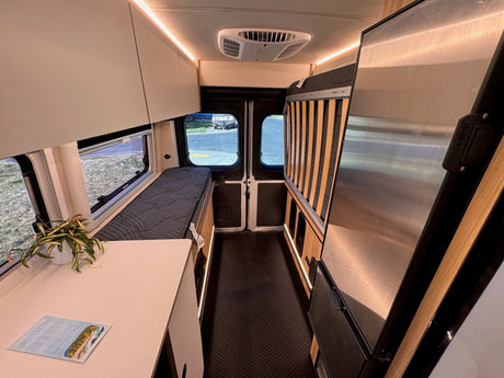 RV rental interior: bed, refrigerator, overhead cabinets, and LED lighting, with rear doors and outdoor view visible.