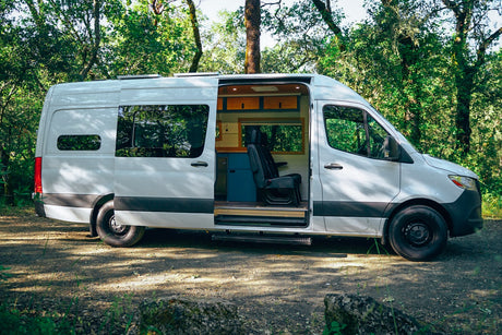 White RV rental camper van, side door open, reveals custom wood and blue interior. Parked in a sunny, green forest setting.