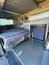 Cozy RV rental interior featuring a made bed with gray bedding, built-in fridge, and diamond-plate floor for your next adventure.